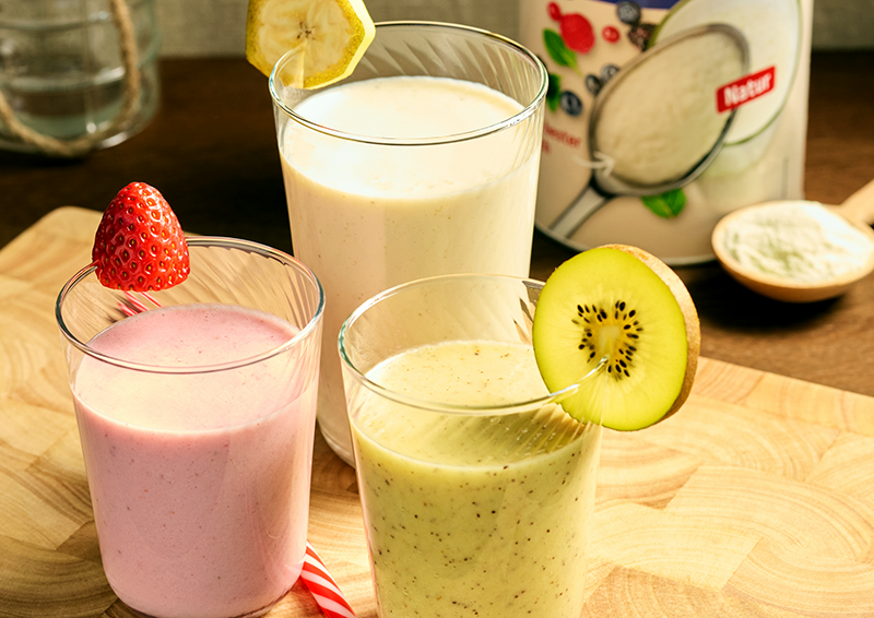 Drei Smoothies in Gläsern mit Erdbeere, Kiwi und Zitrone garniert, daneben Verpackung und Pulver auf Holzbrett