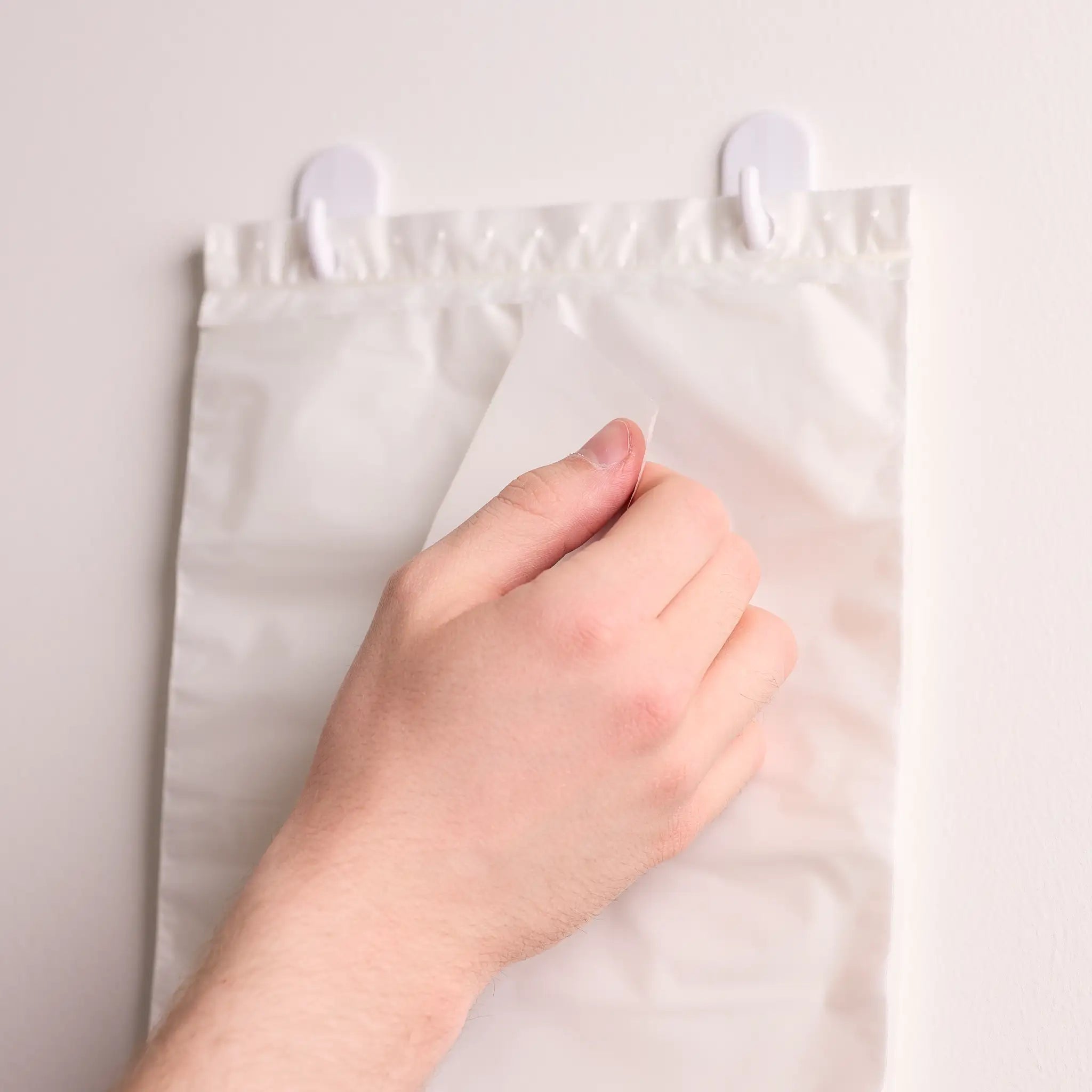 Hand zieht ein Tuch aus einem weißen Beutelspender, der mit Haken an einer hellen Wand befestigt ist.