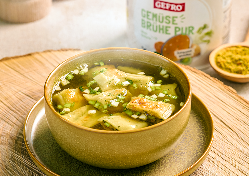 Schüssel mit klarer Gemüsesuppe, gefüllten Teigtaschen und Kräutern, daneben GEFRO Gemüsebrühe Pur im Hintergrund