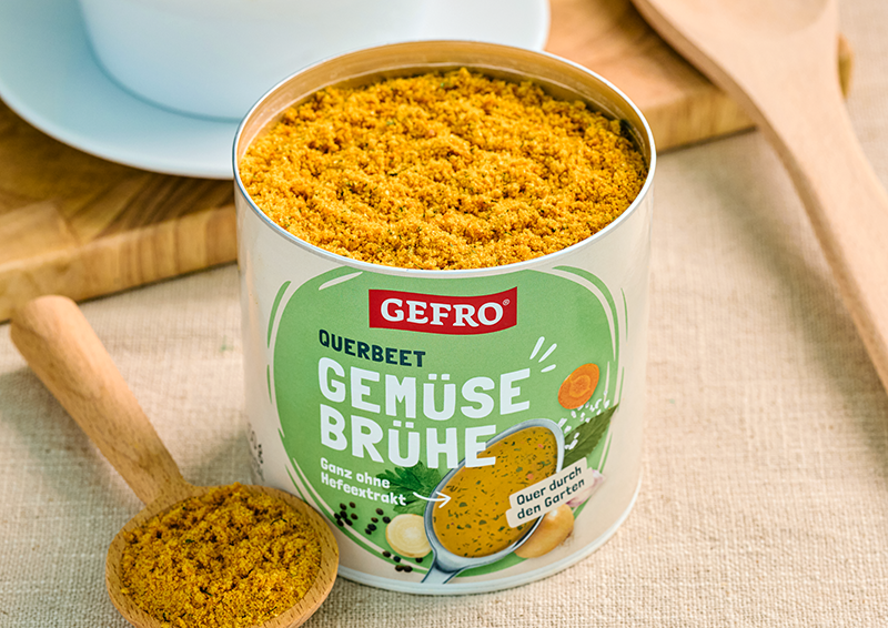 Dose GEFRO Gemüsebrühe mit offenem Pulverinhalt auf Tisch, daneben Holzlöffel mit Brühepulver