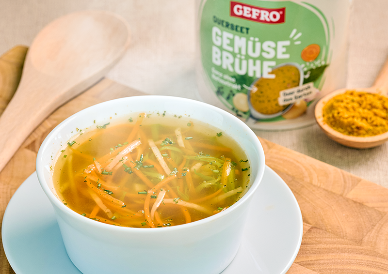 Schüssel mit klarer Gemüsesuppe und Gemüsestreifen auf Untertasse, daneben GEFRO Gemüsebrühe und Holzlöffel
