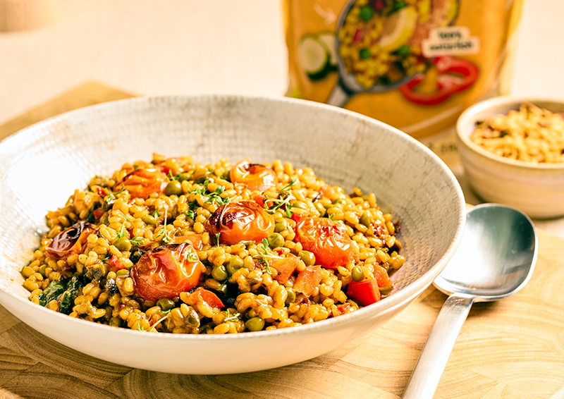 Schüssel mit würzigem Getreidesalat, gerösteten Tomaten und Erbsen auf Holzbrett, daneben Löffel und Verpackung