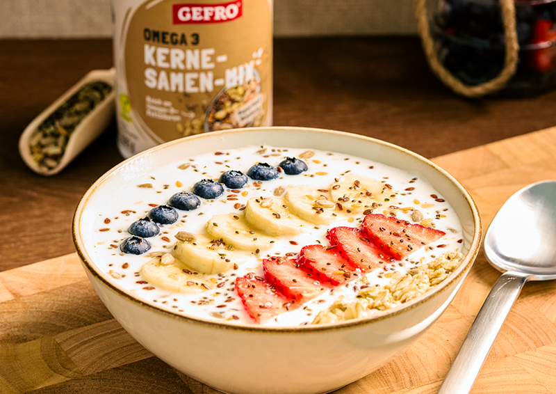 Schale mit Joghurt, Banane, Erdbeeren, Heidelbeeren, Haferflocken und Saaten, daneben Kern-Samen-Mix von GEFRO