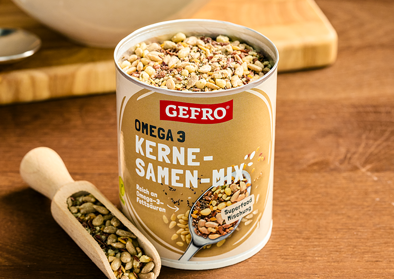 Dose mit GEFRO Omega 3 Kerne-Samen-Mix auf Holztisch, daneben eine Holzschaufel mit Samenmischung.