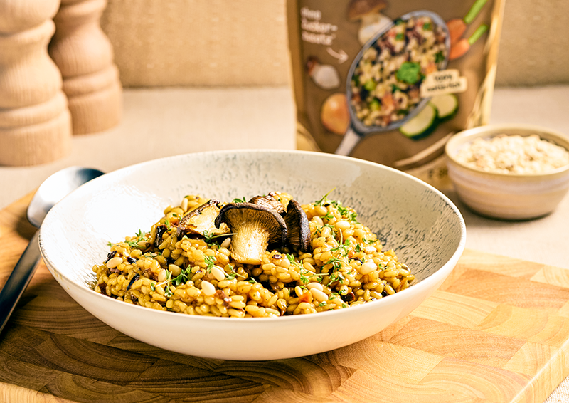 Schale mit cremigem Pilz-Risotto auf Holzbrett, garniert mit Pilzen und Kräutern, Produktverpackung im Hintergrund