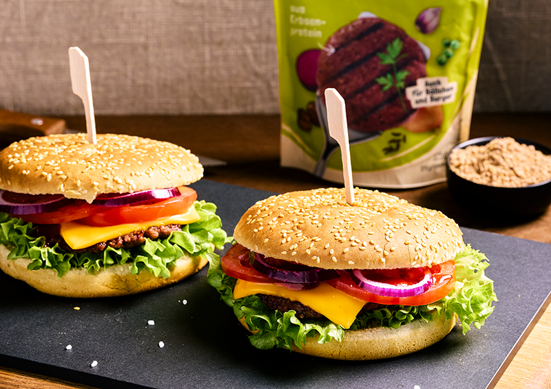 Zwei vegane Burger mit Salat, Käse, Tomate und roten Zwiebeln auf einer Platte, dahinter eine Packung Burger-Mix.