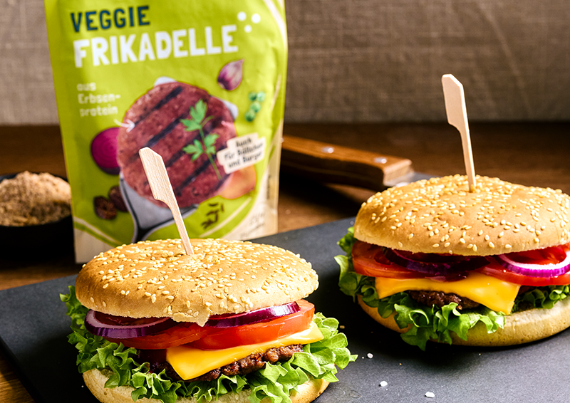 Zwei Burger mit Salat, Tomate, Zwiebeln und Käse vor einer Packung Veggie Frikadelle auf dunkler Platte.
