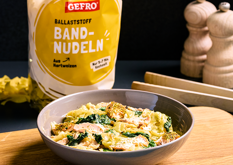 Schüssel mit Bandnudeln, Lachs, Brokkoli und Spinat vor Gefro-Verpackung auf Holzbrett in der Küche