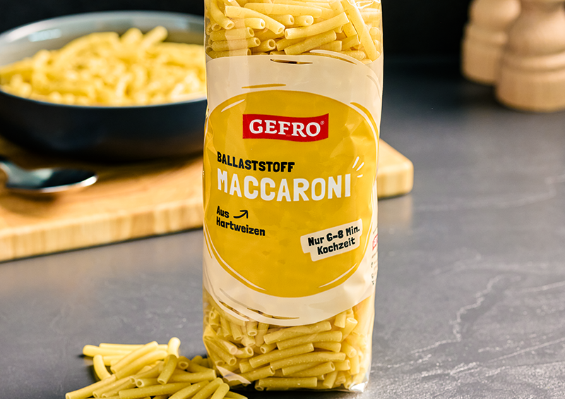 Packung Gefro Ballaststoff-Maccaroni aus Hartweizen auf Küchentisch, mit gekochter Pasta im Hintergrund