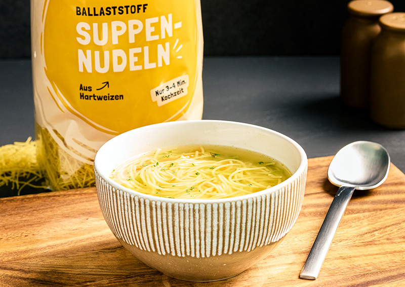 Schüssel mit Nudelsuppe auf Holzbrett, daneben Löffel und Packung Ballaststoff-Suppennudeln aus Hartweizen