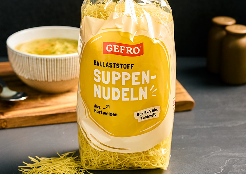 Packung GEFRO Ballaststoff-Suppennudeln aus Hartweizen, dekorativ vor einer Schüssel Suppe auf dem Tisch platziert
