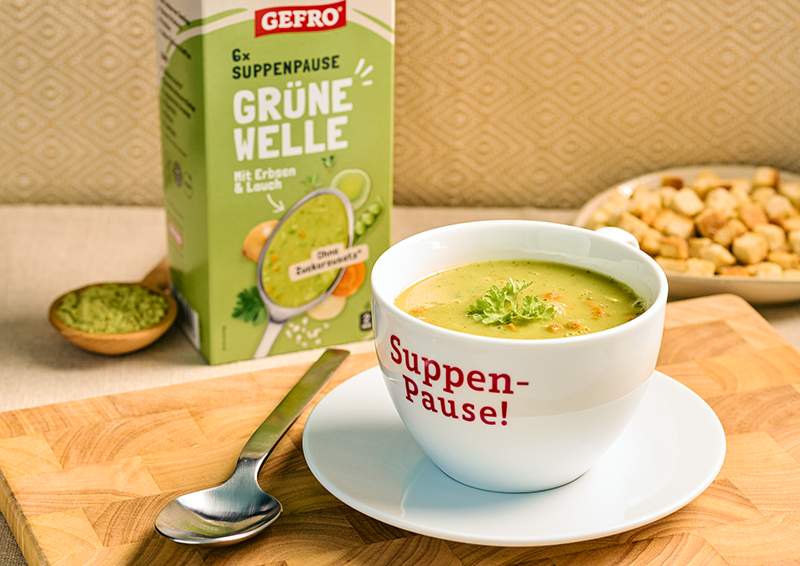 Tasse mit grüner Erbsen-Lauch-Suppe auf Unterteller, daneben GEFRO Verpackung und Löffel auf Holzbrett