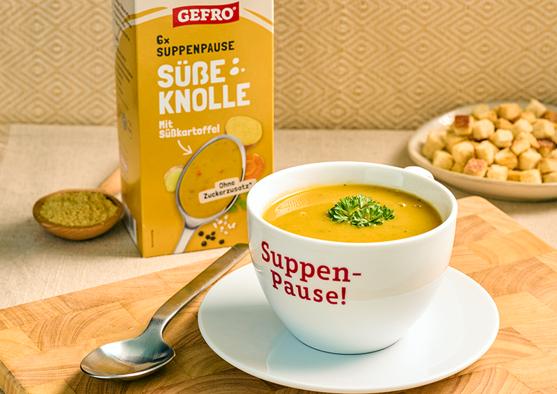 Tasse mit gelber Süßkartoffelsuppe vor Gefro-Verpackung, dazu Löffel und Croutons auf dem Tisch