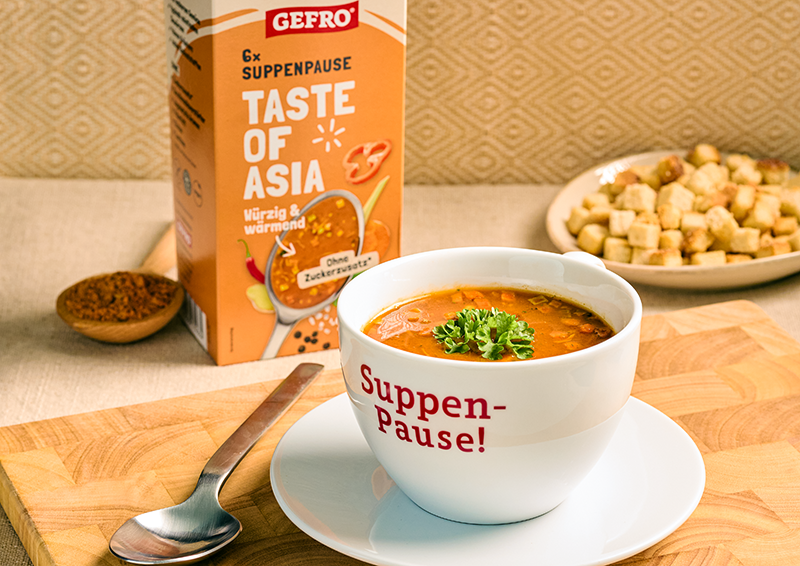 Tasse mit asiatischer Suppe auf Untertasse, daneben GEFRO Suppenpause Taste of Asia und Brotwürfel im Hintergrund