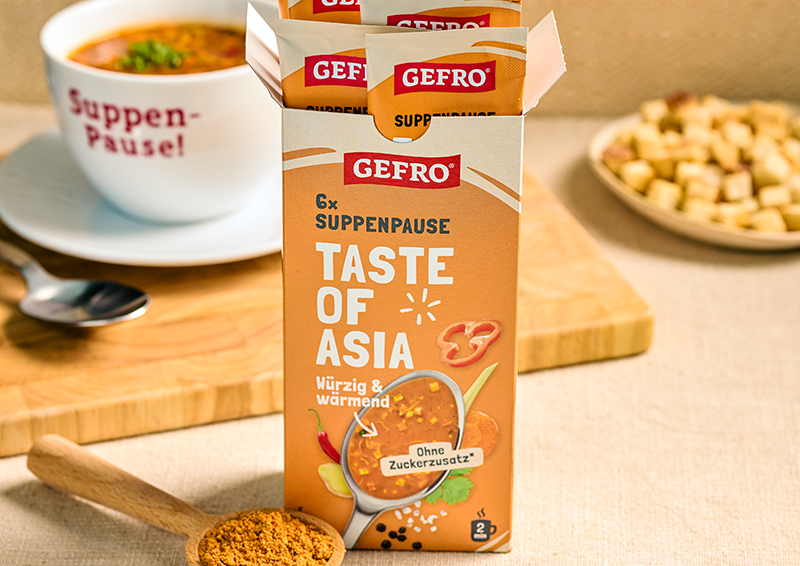 GEFRO Suppenpause Taste of Asia in geöffneter Packung, daneben eine Tasse Suppe und Gewürze auf dem Tisch
