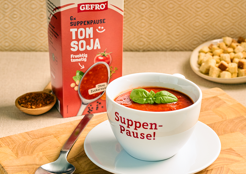 Schale mit Tomaten-Soja-Suppe und Basilikum vor GEFRO-Packung, dazu Löffel und Croûtons im Hintergrund