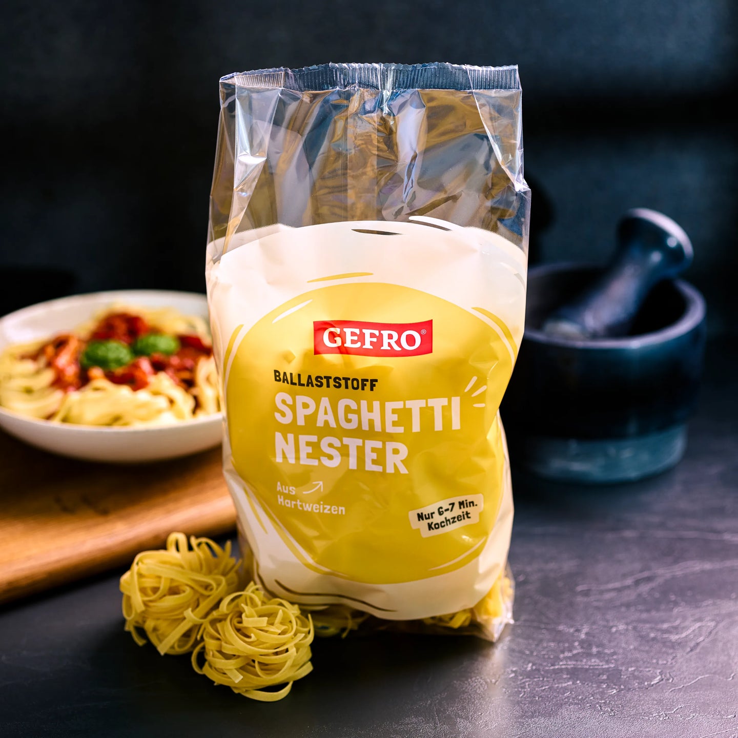 Packung GEFRO Spaghetti-Nester aus Hartweizen, davor lose Nudeln; im Hintergrund ein Teller mit Pasta.