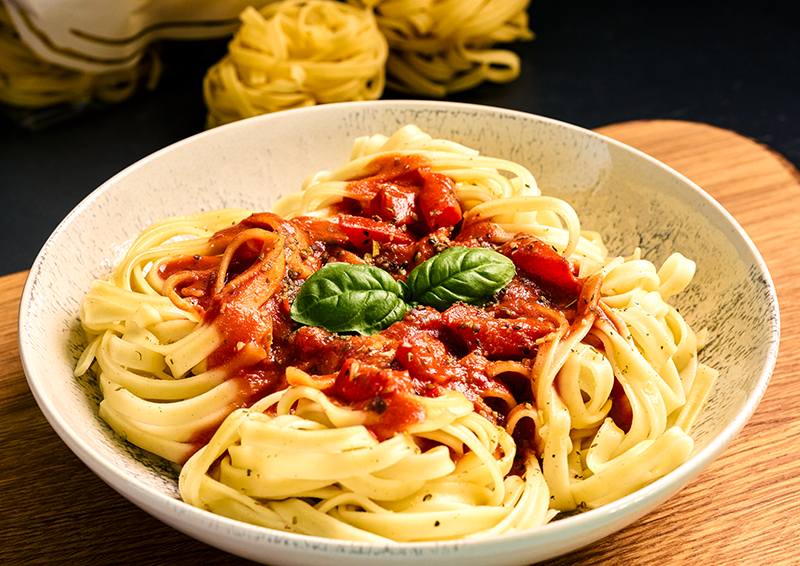 Teller mit Tagliatelle in Tomatensauce, garniert mit frischem Basilikum, servierfertig auf Holzbrett präsentiert