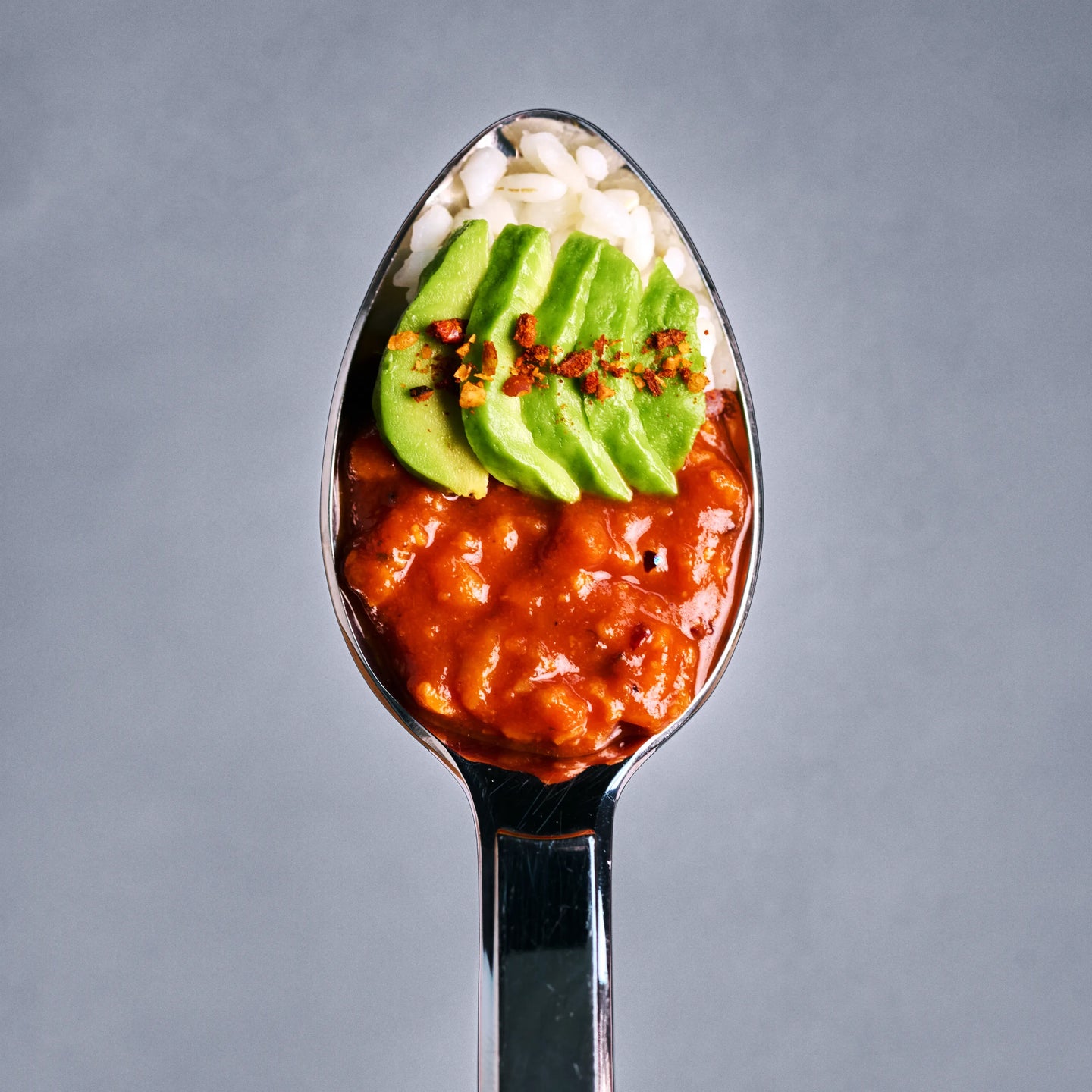 Löffel mit Reis, roter Sauce und Avocadoscheiben, von oben fotografiert auf schlichtem grauem Hintergrund