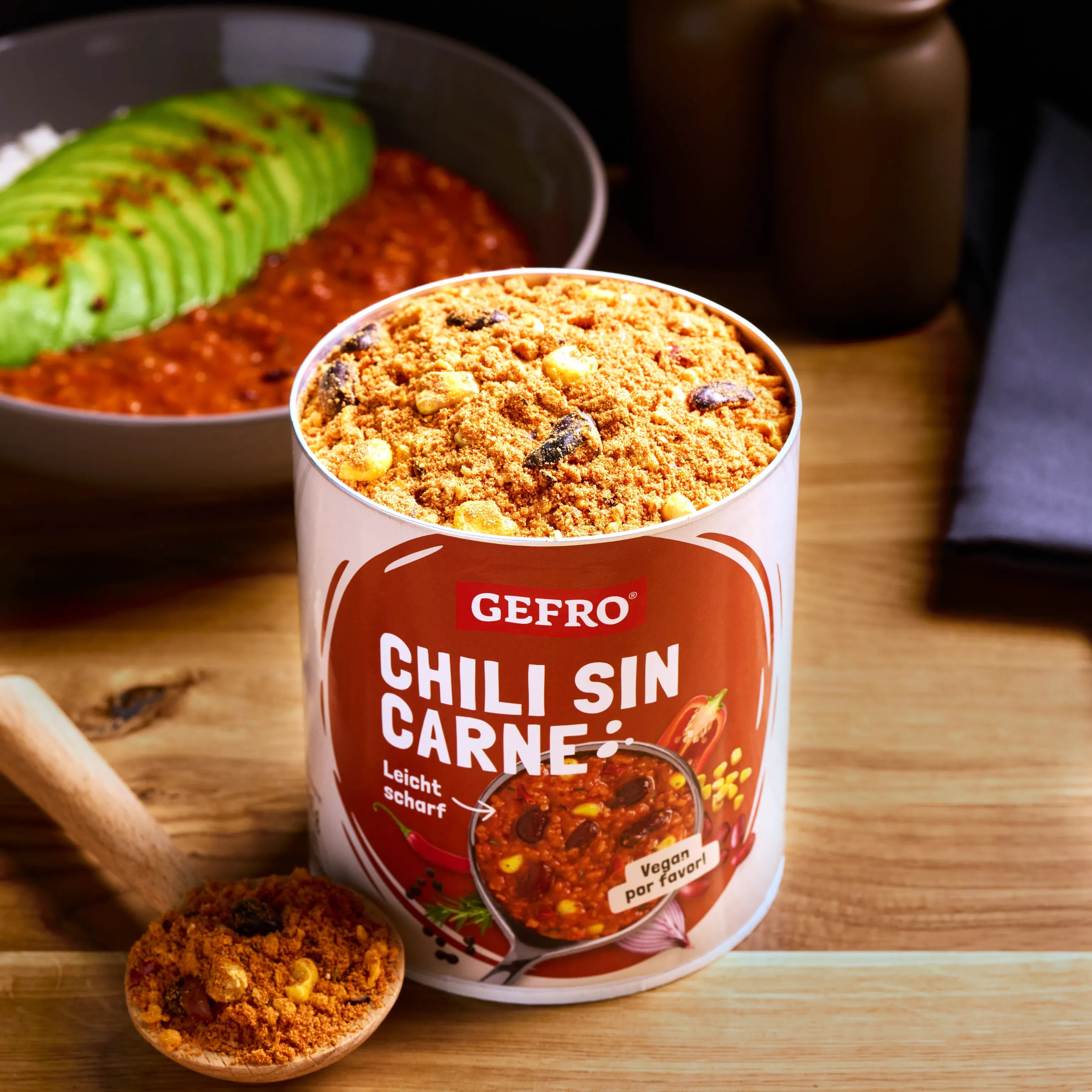 Dose GEFFRO Chili sin Carne mit Gewürzmischung auf Holztisch, daneben Löffel und servierte Chili-Bowl mit Avocado