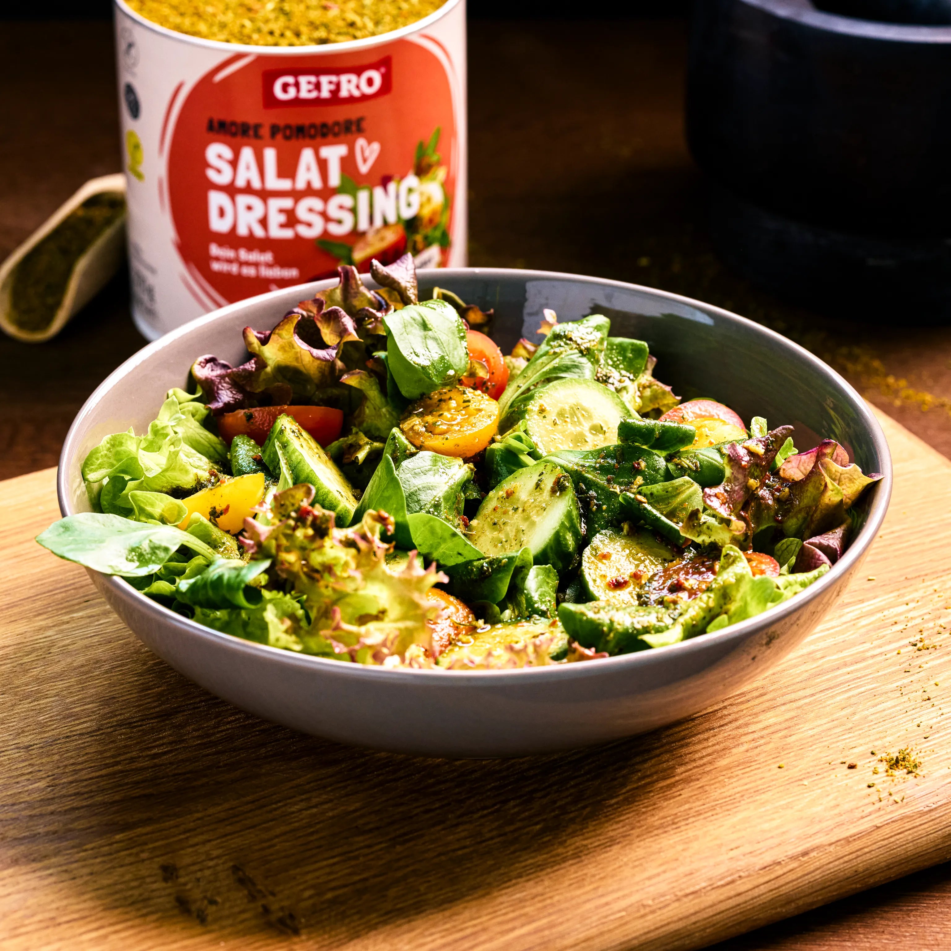 Schüssel mit frischem gemischtem Salat und Gurke auf Holzbrett, daneben eine Dose Gefro Salat-Dressing Tomate