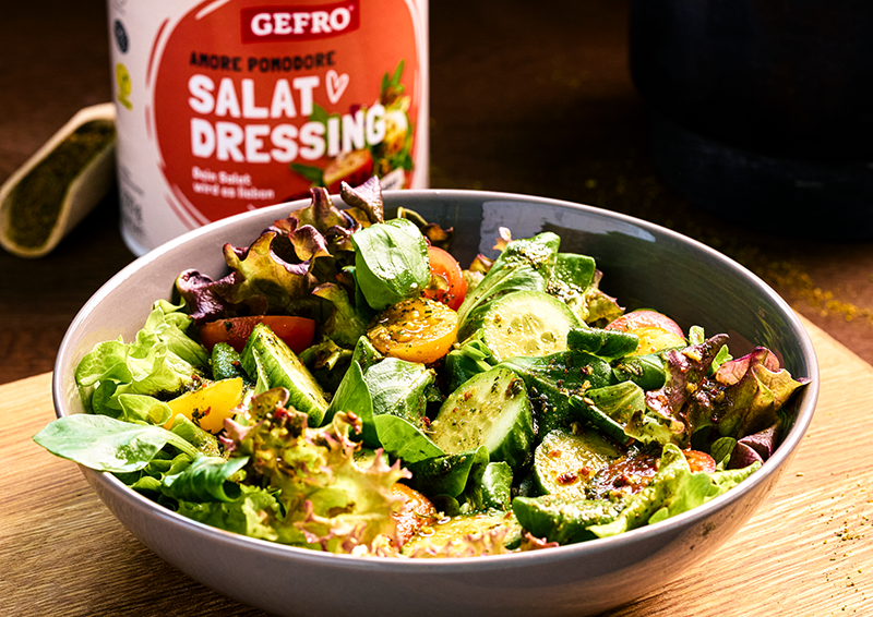 Schüssel mit frischem gemischtem Salat, Gurke und Tomaten, davor GEFRO Salat Dressing auf Holztisch