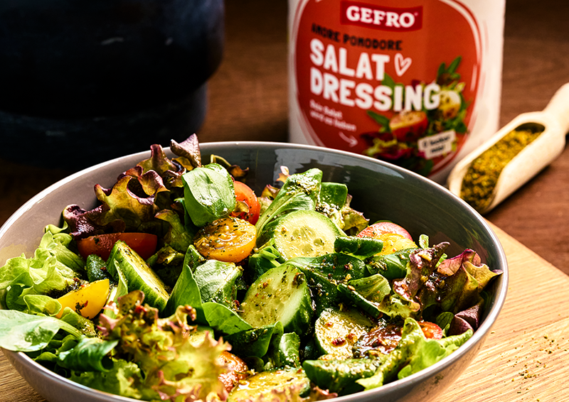 Schüssel mit frischem Blattsalat, Gurken und Tomaten, daneben Salatdressing von Gefro und Gewürze auf Holzbrett