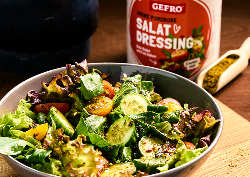 Schüssel mit frischem gemischtem Salat und Gurke, dazu Salatdressing im Hintergrund auf einem Holztisch