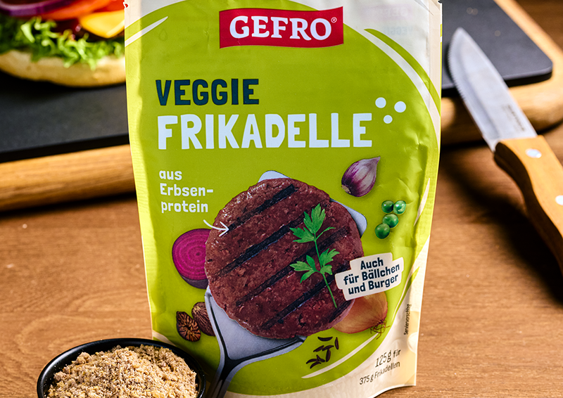 Verpackung von Gefro Veggie Frikadelle aus Erbsenprotein auf Holztisch, daneben Messer und Schale mit Gewürz