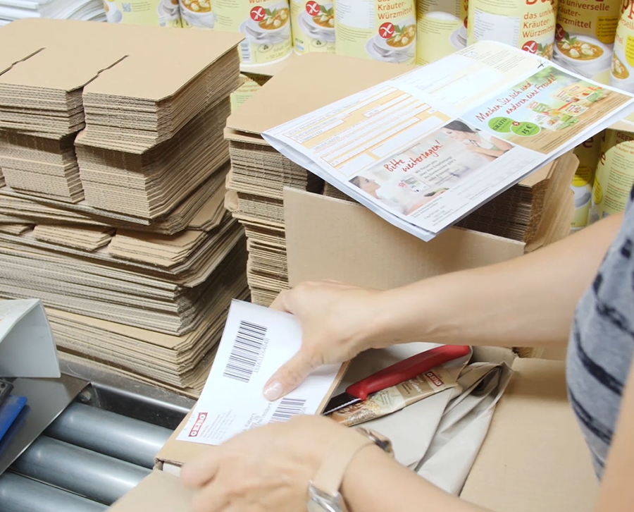 Mitarbeiterin verpackt Ware in Karton, daneben liegen Versandetikett, Flyer und gestapelte Verpackungen.