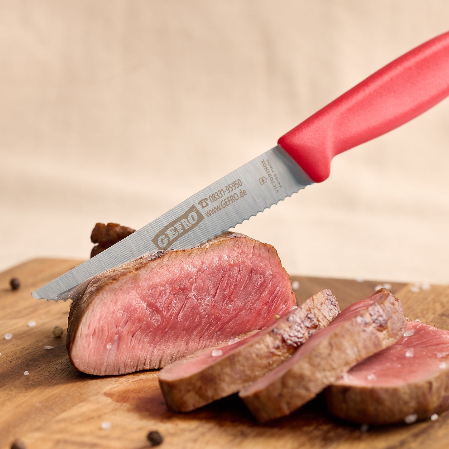 Rotes Messer mit Wellenschliff schneidet rosa gebratenes Fleisch auf einem Holzbrett in feine Scheiben.