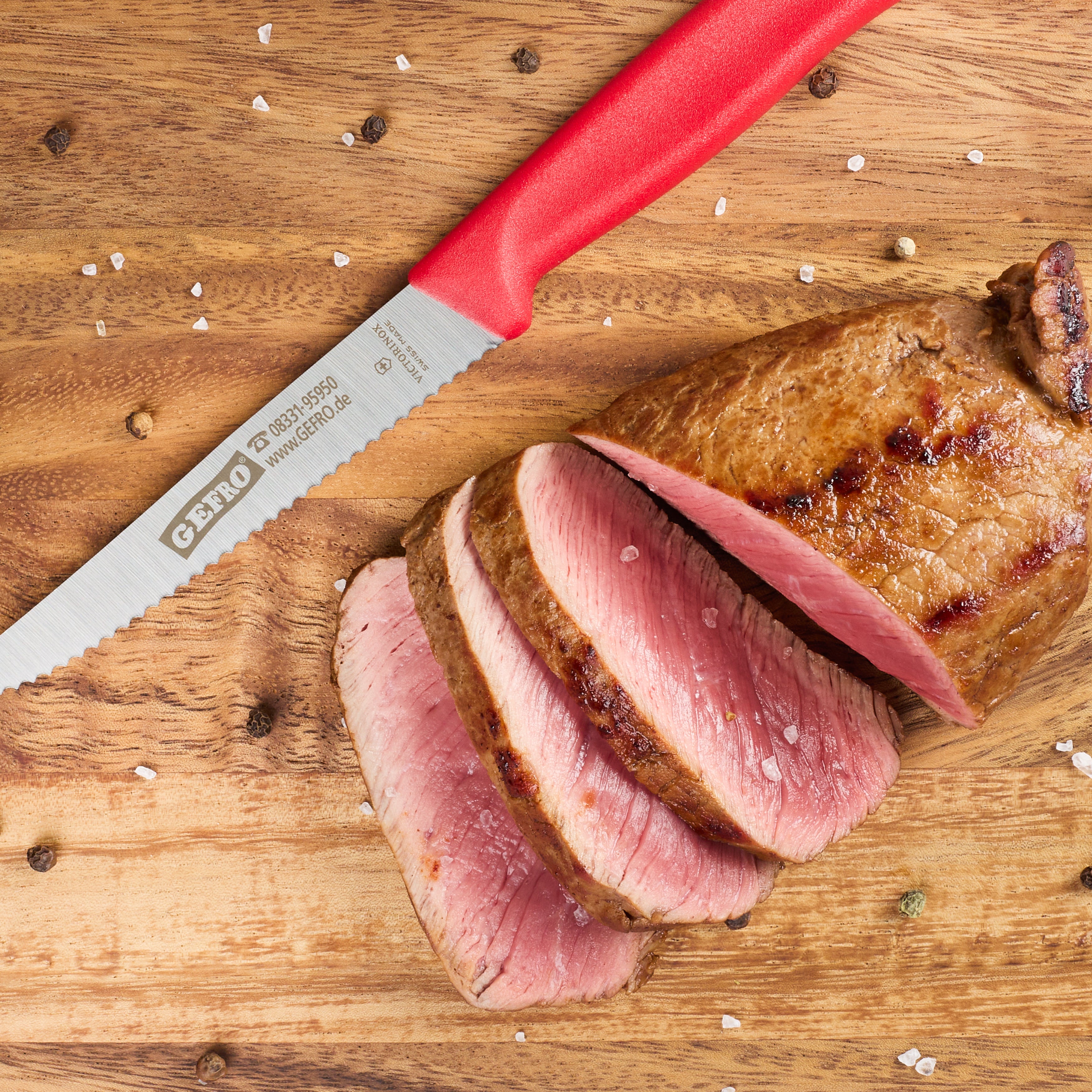 Gezahntes Küchenmesser mit rotem Griff neben aufgeschnittenem rosa gebratenem Fleisch auf Holzbrett mit Gewürzen