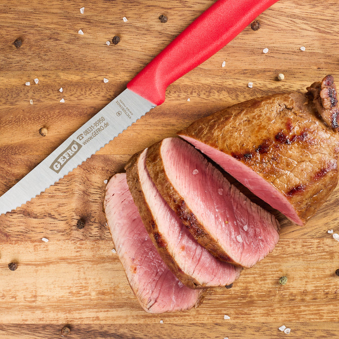 Gezahntes Küchenmesser mit rotem Griff neben aufgeschnittenem rosa gebratenem Fleisch auf Holzbrett mit Gewürzen