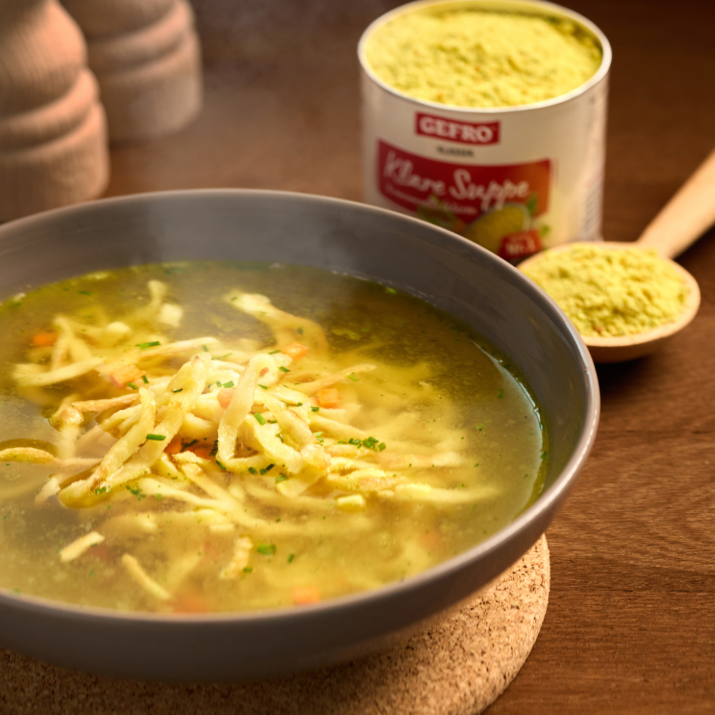 Schüssel mit klarer Suppe und feinen Streifen, dahinter Becher mit Klare Suppe und Löffel mit Pulver auf Holztisch