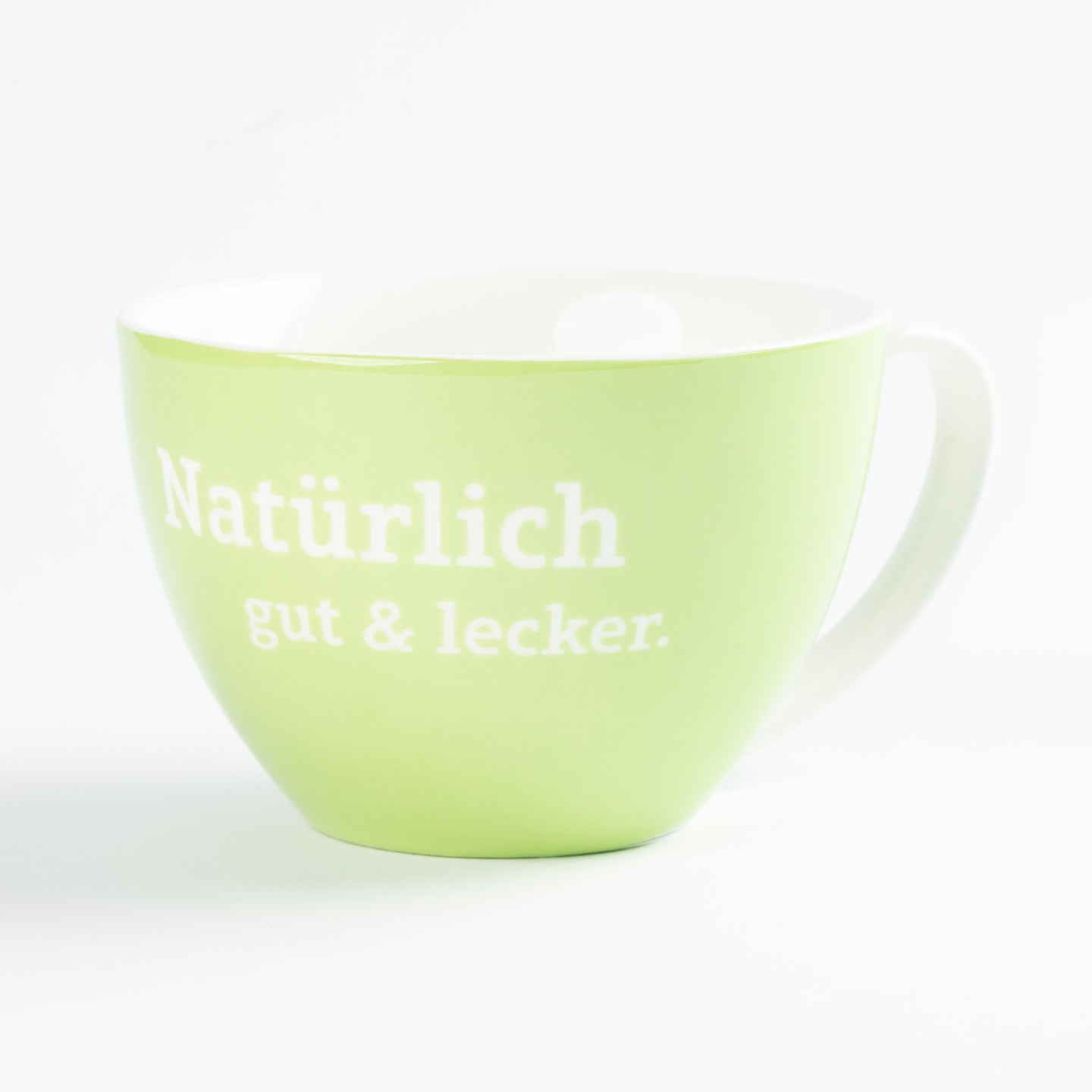 Grüne Kaffeetasse mit weißem Schriftzug „Natürlich gut & lecker“ auf hellem Hintergrund, Keramikbecher.