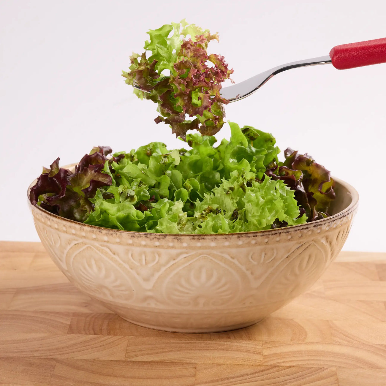 Keramikschüssel mit gemischtem Blattsalat auf Holztisch, darüber eine Gabel mit Salat zum Servieren.