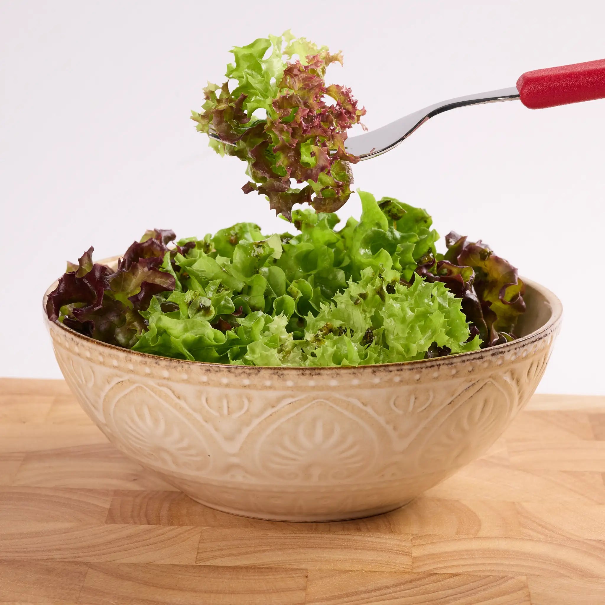 Keramikschüssel mit gemischtem Blattsalat auf Holztisch, darüber eine Gabel mit Salat zum Servieren.
