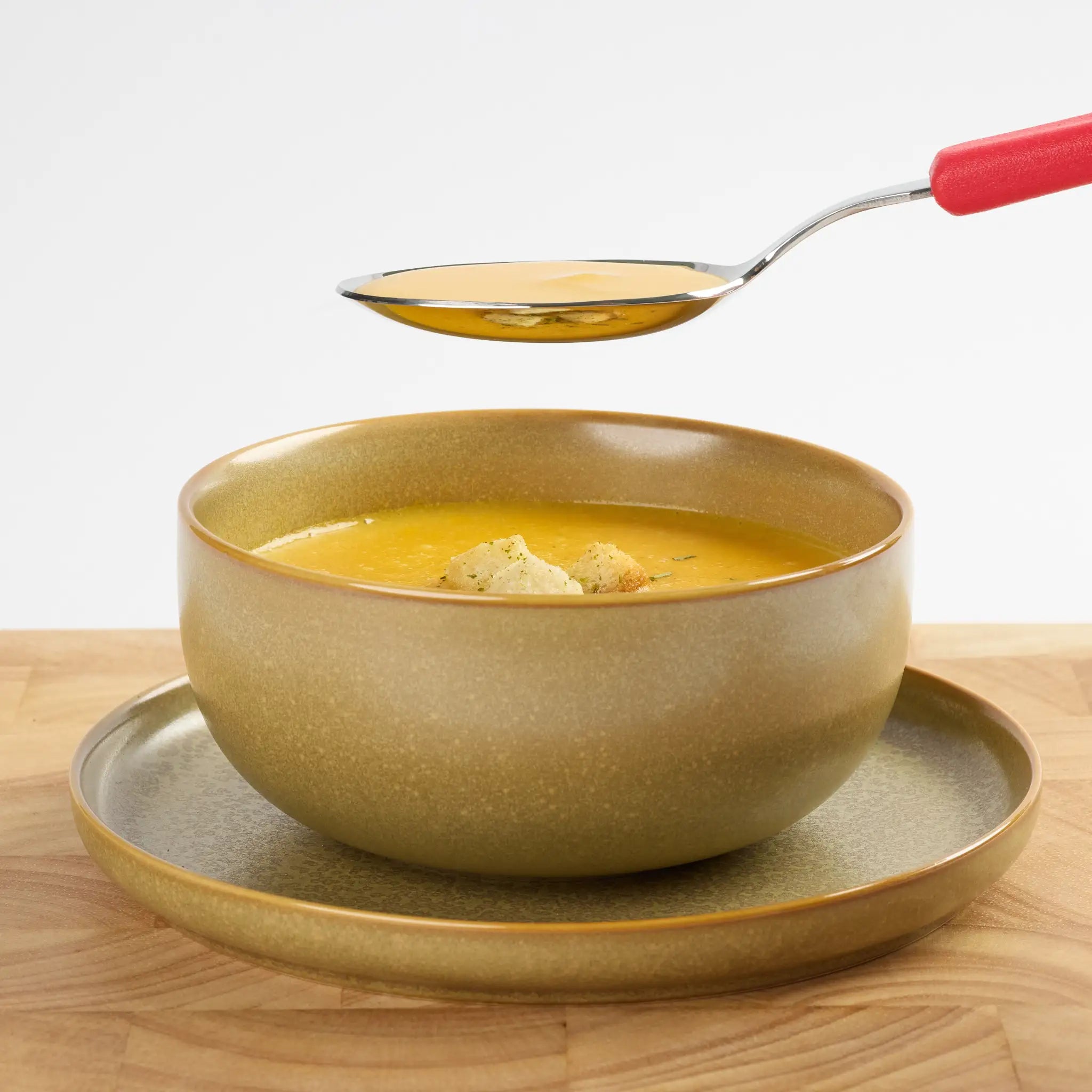 Goldene Schale mit cremiger Suppe auf passendem Teller, darüber ein Löffel mit rotem Griff beim Servieren