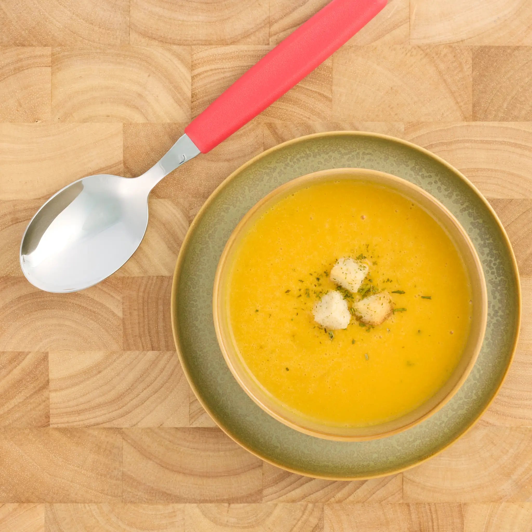 Schale mit gelber Cremesuppe auf grünem Teller, daneben ein großer Löffel mit rotem Griff auf Holzbrett.