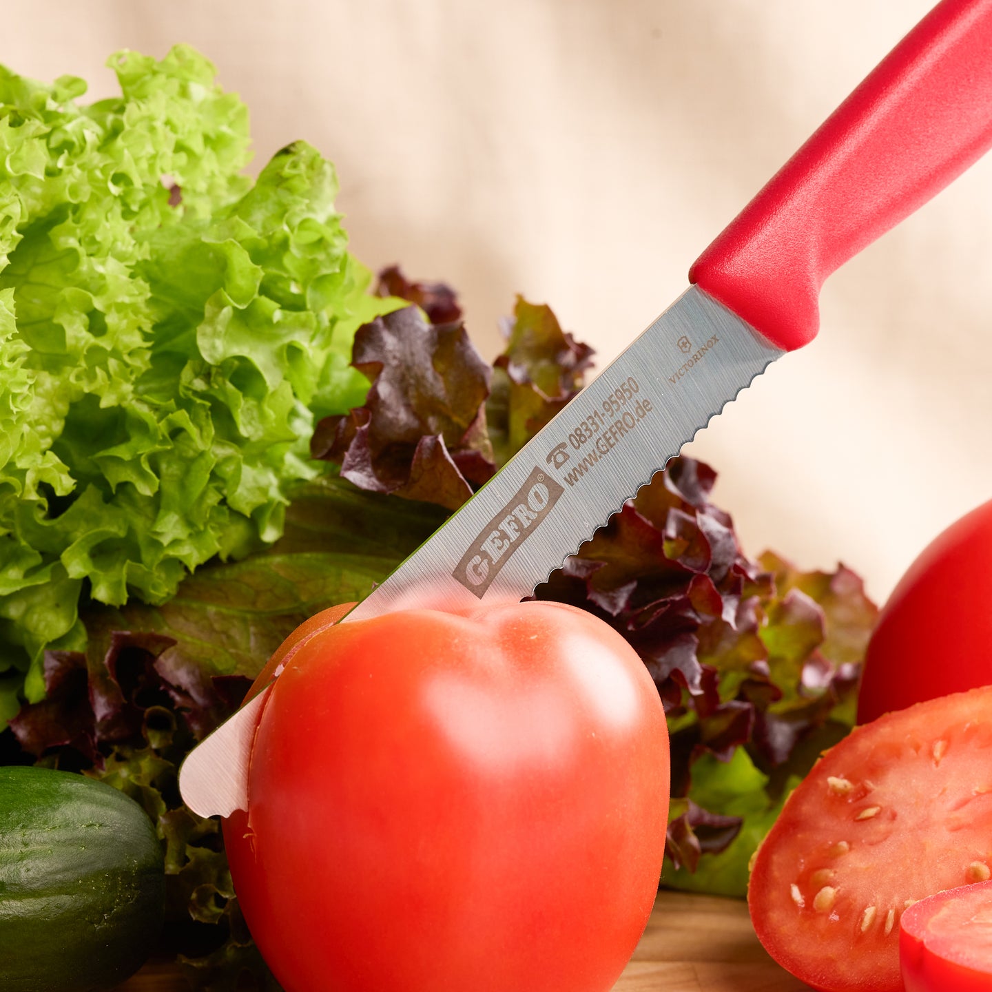 Rotes Gemüsemesser mit Wellenschliff schneidet eine Tomate, umgeben von Salat und weiterem frischem Gemüse.