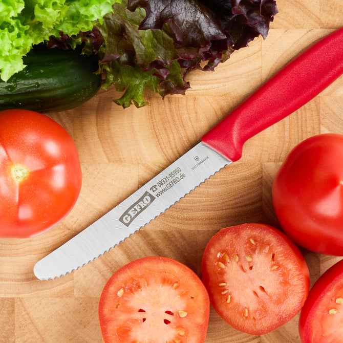 Rotes Tomatenmesser mit Wellenschliff auf Holzbrett, umgeben von Tomaten, Salat und Gurke in der Küche