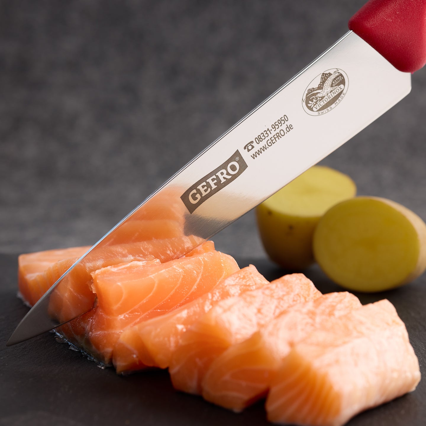 Scharfes Küchenmesser mit rotem Griff schneidet frischen Lachs auf dunkler Unterlage, Kartoffeln im Hintergrund