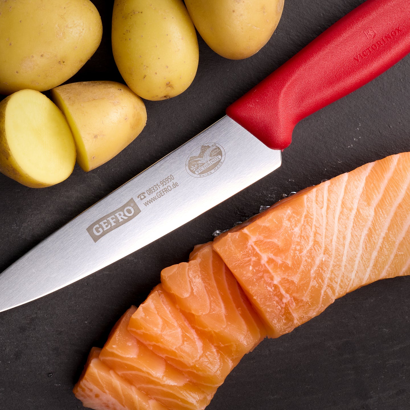 Küchenmesser mit rotem Griff neben geschnittenem Lachs und Kartoffeln auf dunkler Fläche, für präzises Schneiden
