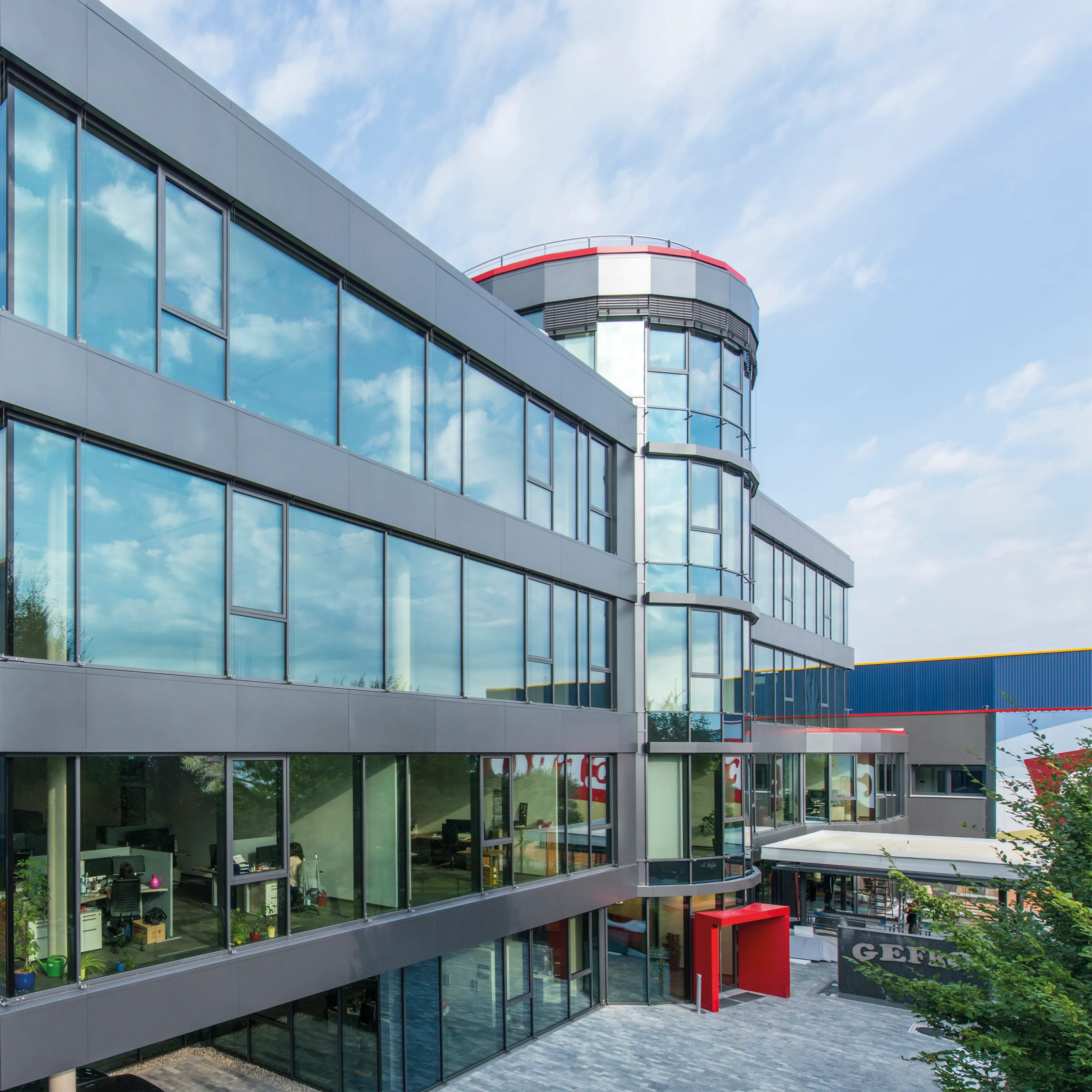 Modernes Bürogebäude mit großer Glasfassade, abgerundetem Eckturm und rotem Eingang bei hellem Himmel