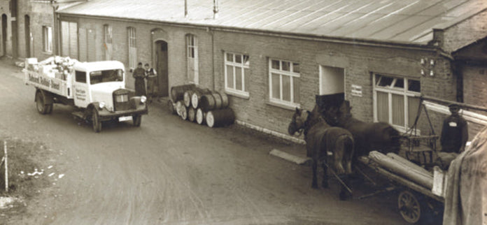 Historische Hofszene mit Lastwagen, Fässern und Pferdewagen vor einem Backsteingebäude in Sepiatönen