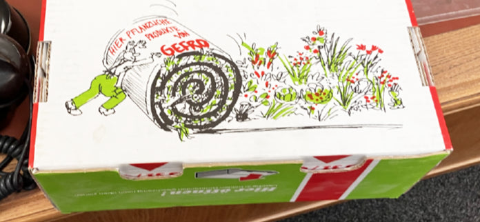 Grün-weiße Produktverpackung mit Zeichnung einer ausgerollten Pflanzenmatte und Blumenwiese auf einem Tisch