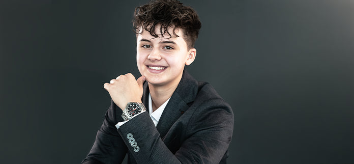 Lächelnde Person im dunklen Blazer präsentiert eine Armbanduhr am Handgelenk vor grauem Hintergrund.