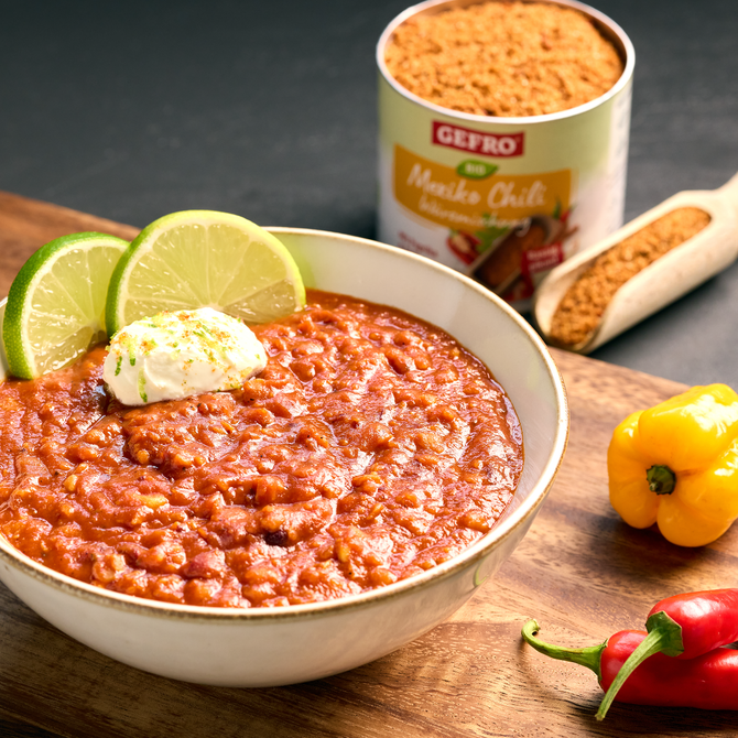 Schüssel mit rotem Chili con Carne, garniert mit Limetten und Creme, daneben Gewürzdose und frische Paprika