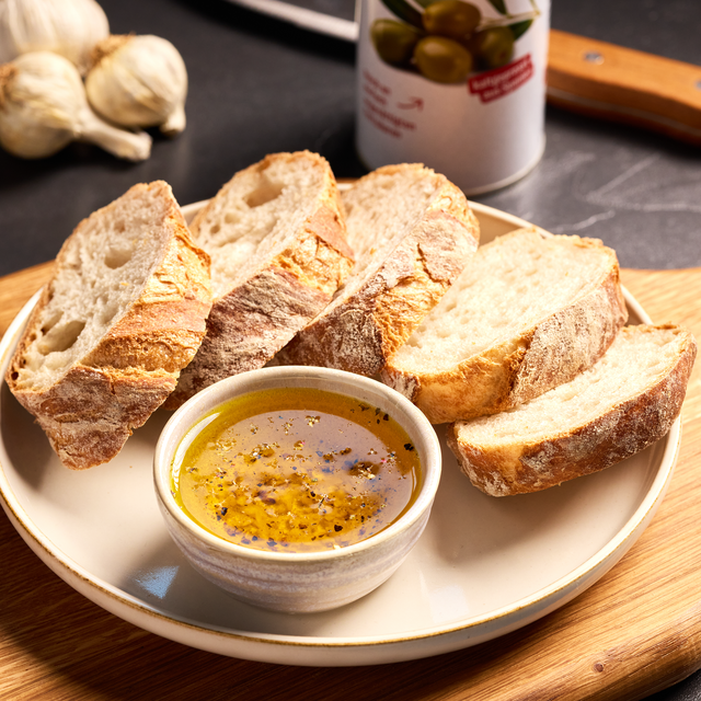 Aufgeschnittenes Weißbrot auf einem Teller mit Schale aus Olivenöl und Gewürzen, daneben Knoblauch und Olivendose.