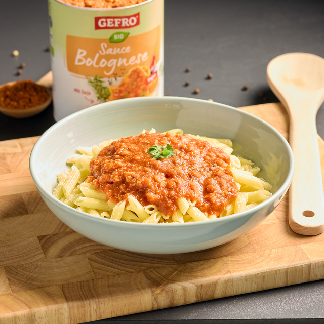 Schale mit Penne und Bolognese-Sauce auf Holzbrett, dahinter Dose von Gefro Bio Sauce Bolognese und Kochlöffel